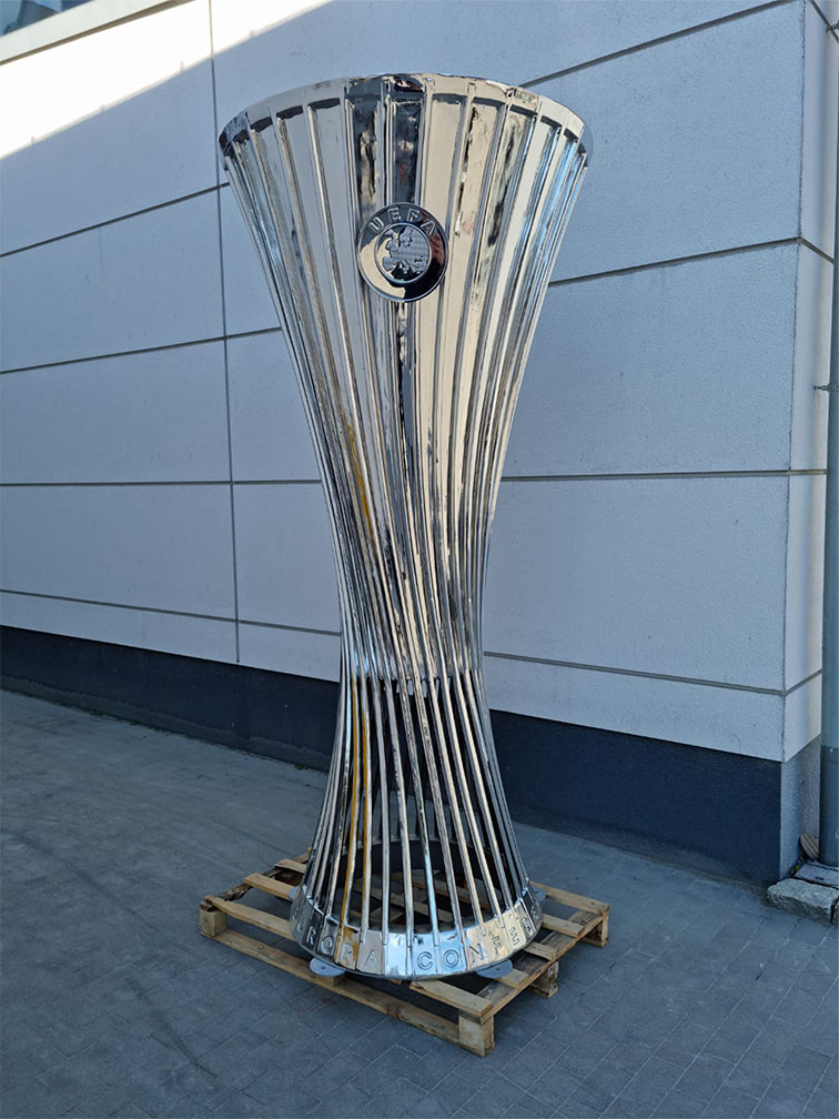 Srebrny puchar UEFA o smukłym, rozszerzającym się ku górze kształcie, stojący na drewnianej palecie na tle jasnoszarej ściany z paneli.