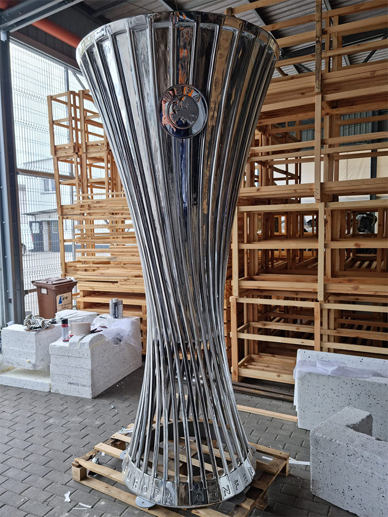 Duży srebrny puchar UEFA Europa Conference League stojący na drewnianej palecie w magazynie z drewnianymi regałami i pudełkami w tle