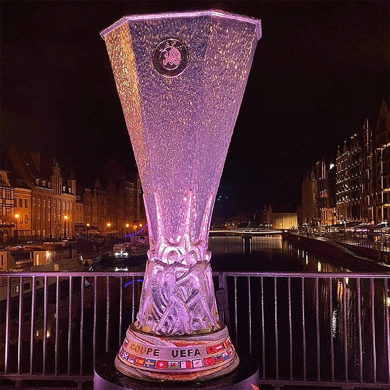 Podświetlony na fioletowo puchar UEFA ustawiony na tle nocnego miasta z oświetlonymi budynkami i kanałem