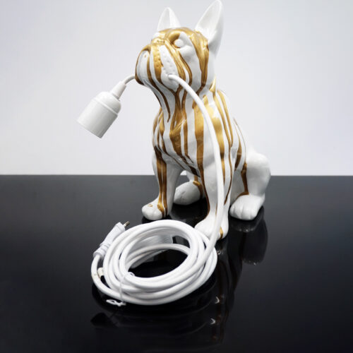 Biało-złoty ceramiczny lampion w kształcie psa trzymający w pysku biały kabel elektryczny na czarnej powierzchni.