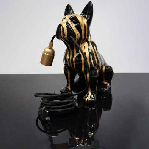 Figurka czarnego psa z pozłacanymi paskami trzymająca w pysku złotą oprawkę na żarówkę z czarnym kablem elektrycznym leżącym na błyszczącej czarnej powierzchni
