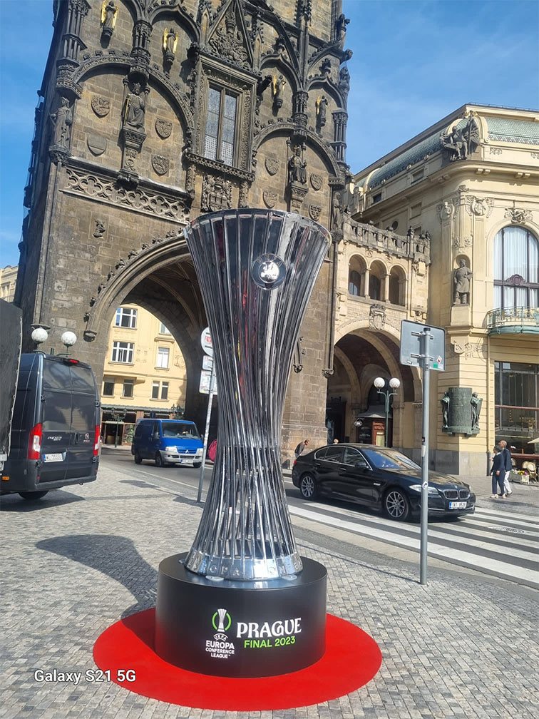 Replika pucharu Liga Konferencji Europy UEFA 2023 na czarnym cokole z napisem PRAGUE FINAL 2023 wystawiona na kamiennym placu z czerwonym dywanikiem, z zabytkową gotycką bramą i klasyczną architekturą w tle, kilka samochodów i przechodniów wokół