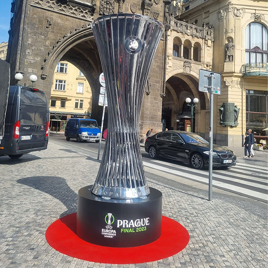 Srebrna statuetka trofeum na czarnym podstawie z napisem PRAGUE FINAL 2023 i logo Europa Conference League, ustawiona na czerwonym okręgu na bruku w tle zabytkowy łuk i budynki, a także przejeżdżające samochody i ludzie na przejściu dla pieszych.