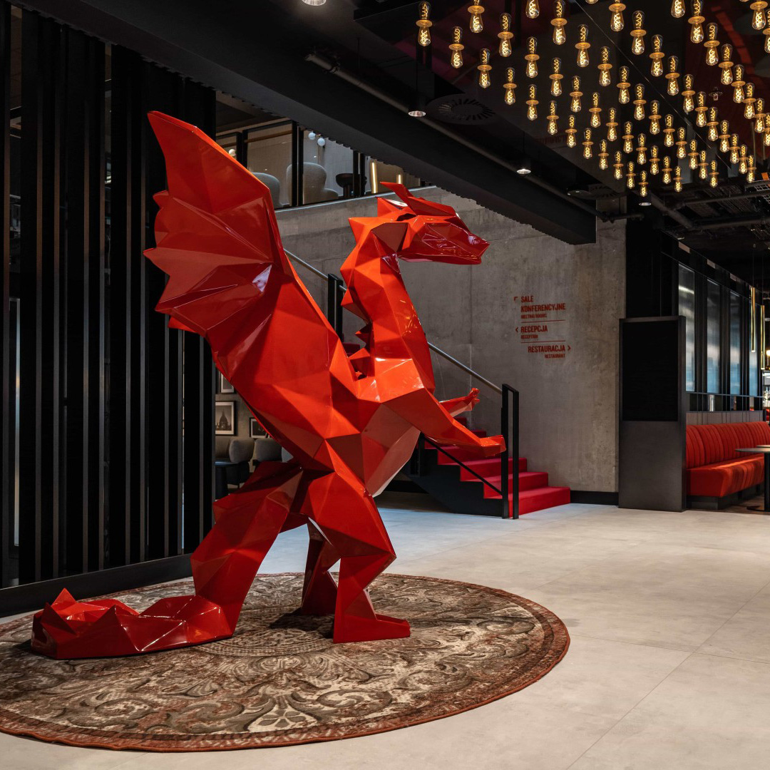 Czerwona geometryczna rzeźba smoka ze skrzydłami stojąca na okrągłym dywanie w nowoczesnym wnętrzu z czerwonymi schodami i wiszącymi żarówkami na suficie.