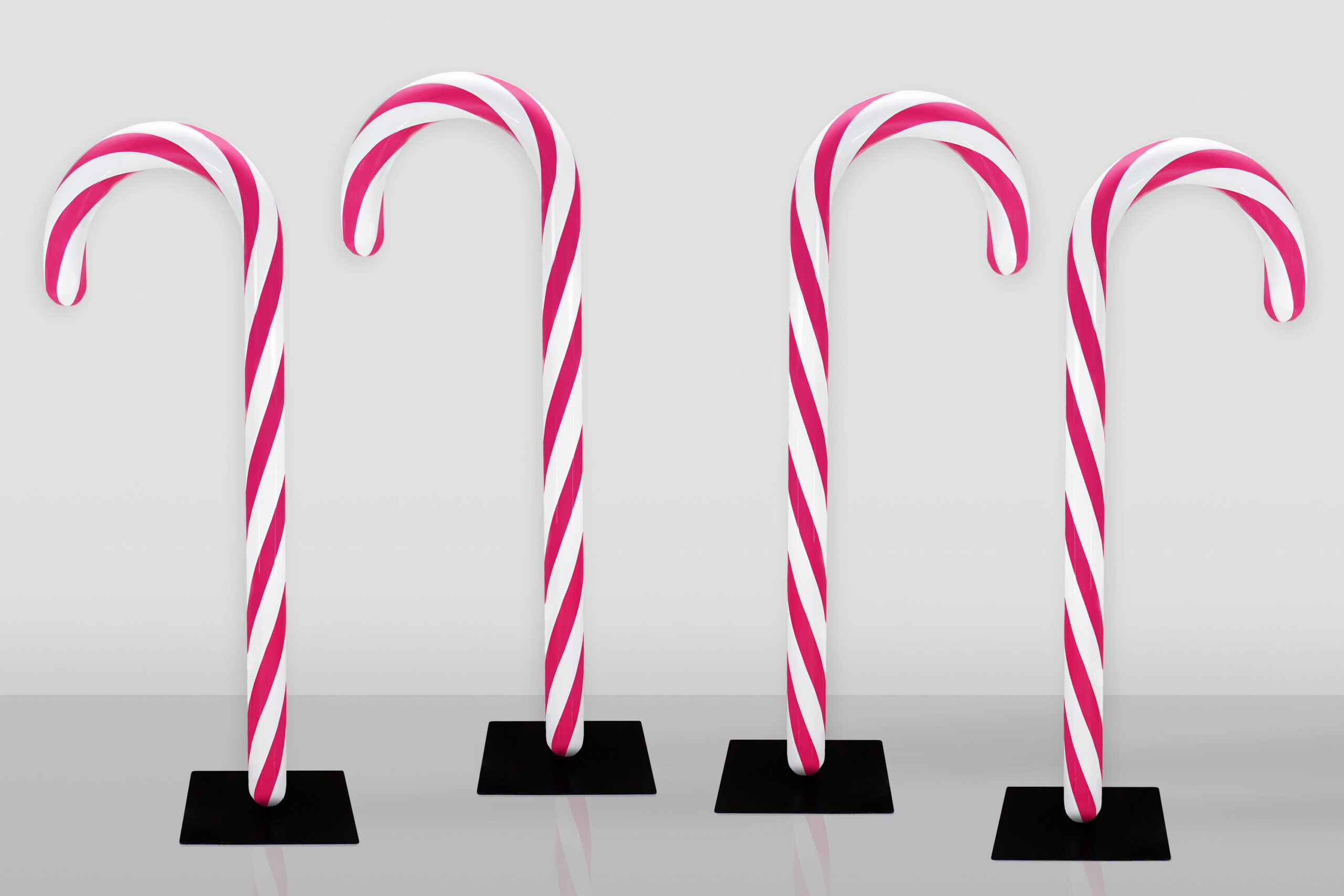 Figura Dekoracyjna Cukrowa Laska Świąteczna Na Podstawie 3 Cztery duże lizaki w kształcie laski z czerwono-białymi spiralnymi paskami stojące na czarnych podstawach na szarej powierzchni przed jasnoszarym tłem.
