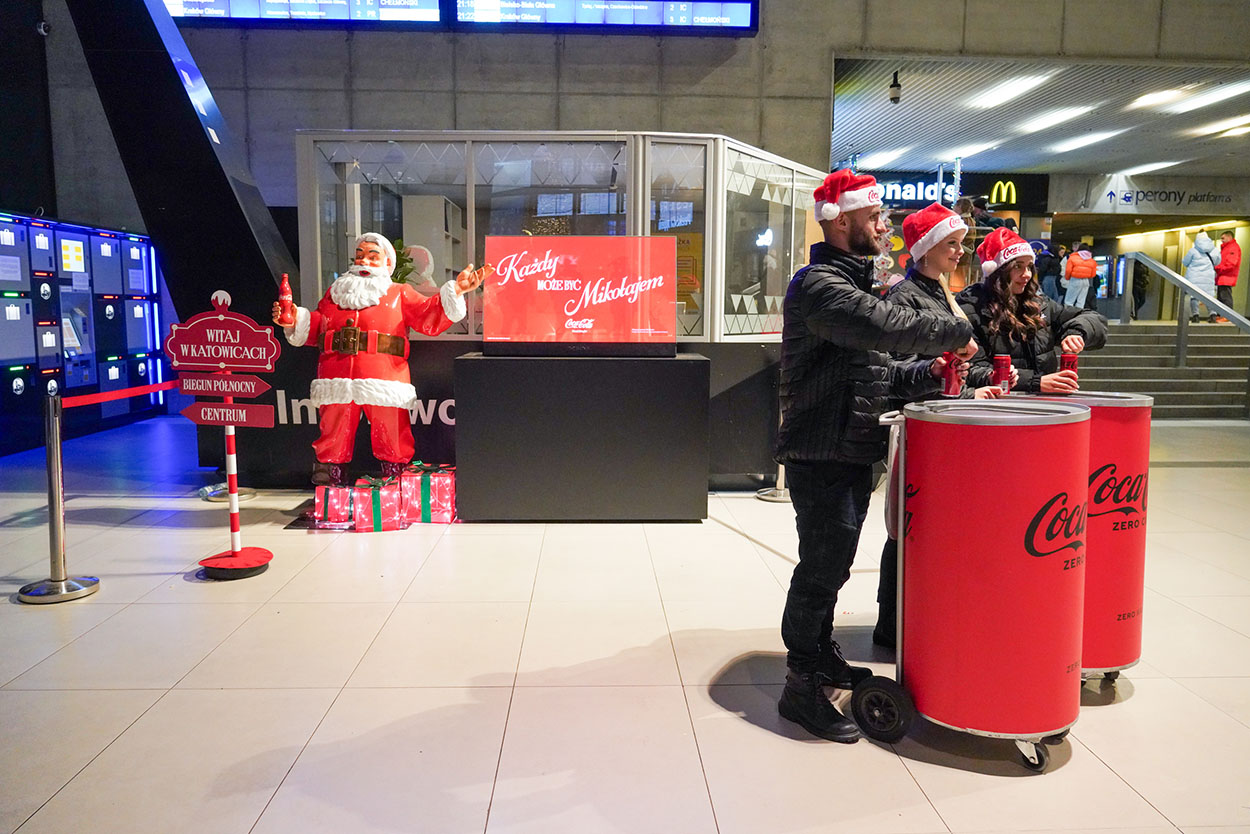 Trzy osoby w czapkach św. Mikołaja stoją przy czerwonych pojemnikach Coca-Coli w hali dworca, obok stoi figura św. Mikołaja z butelką Coca-Coli i znakiem 