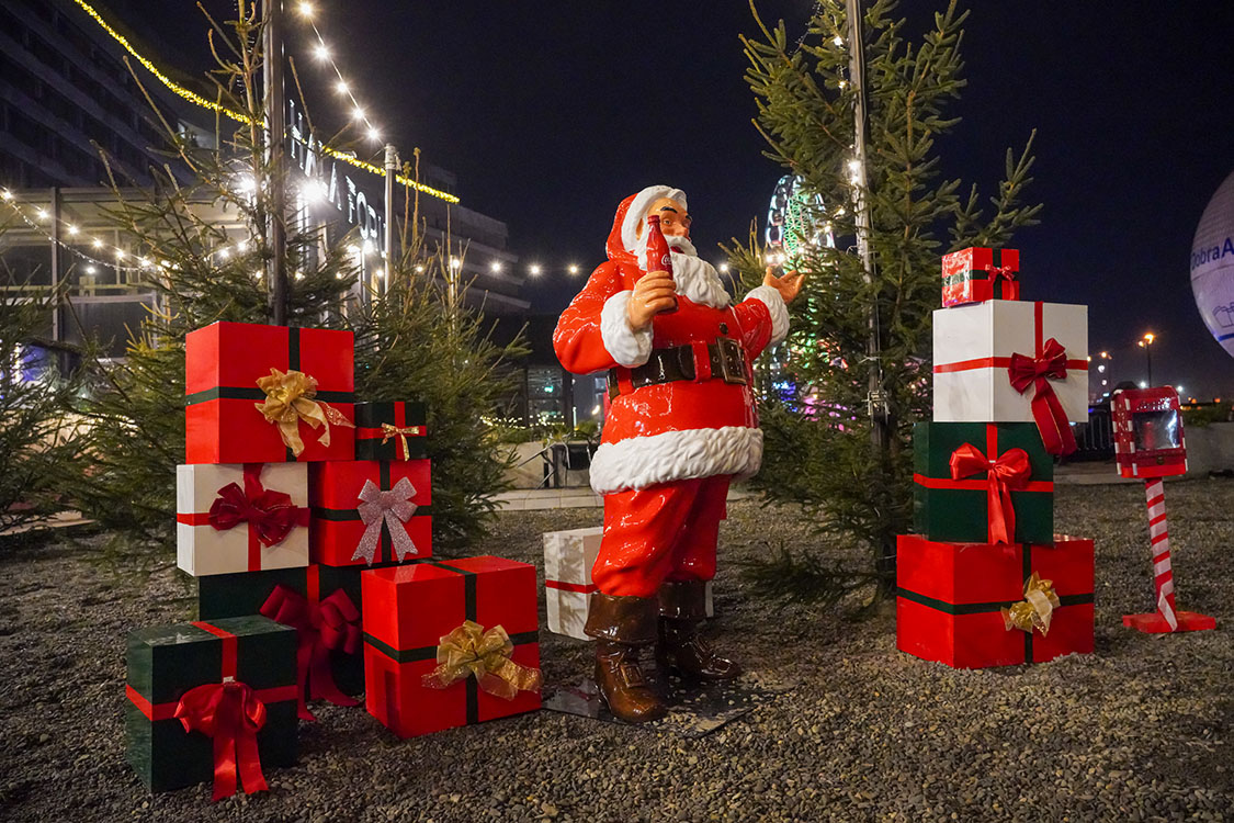 Figurka Mikołaja trzymającego butelkę napoju w otoczeniu świątecznie udekorowanych prezentów i małych drzewek iglastych oświetlonych lampkami nocą w plenerze.