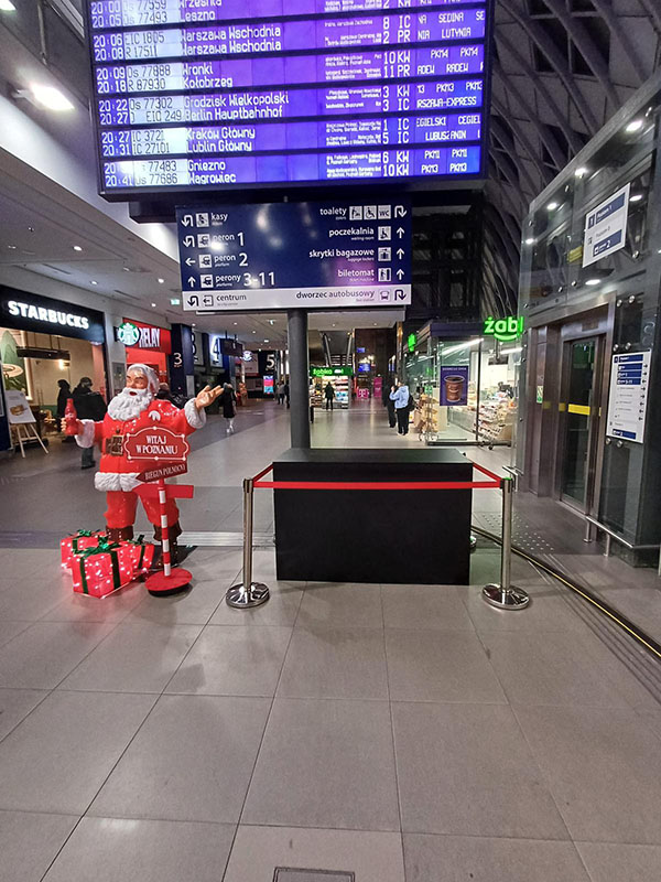 Wnętrze dworca z tablicą odjazdów pociągów na górze, na dole figura Mikołaja z prezentami i napisem Witaj w Poznaniu oraz sklepami Starbucks i Żabka w tle.