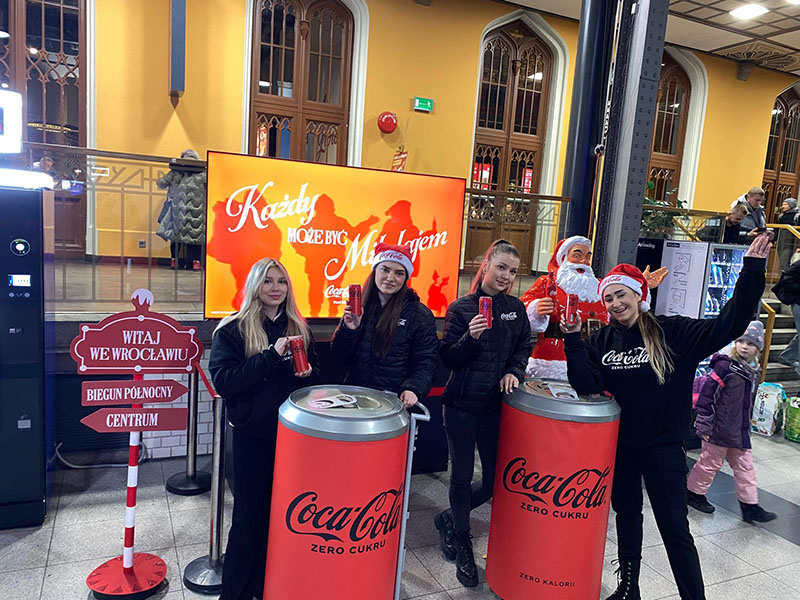 Cztery młode kobiety w ciemnych kurtkach i czapkach Mikołaja pozują z puszkami Coca-Coli przy dwóch dużych stojakach w kształcie puszek Coca-Coli Zero Cukru, na tle ekranu z napisem 