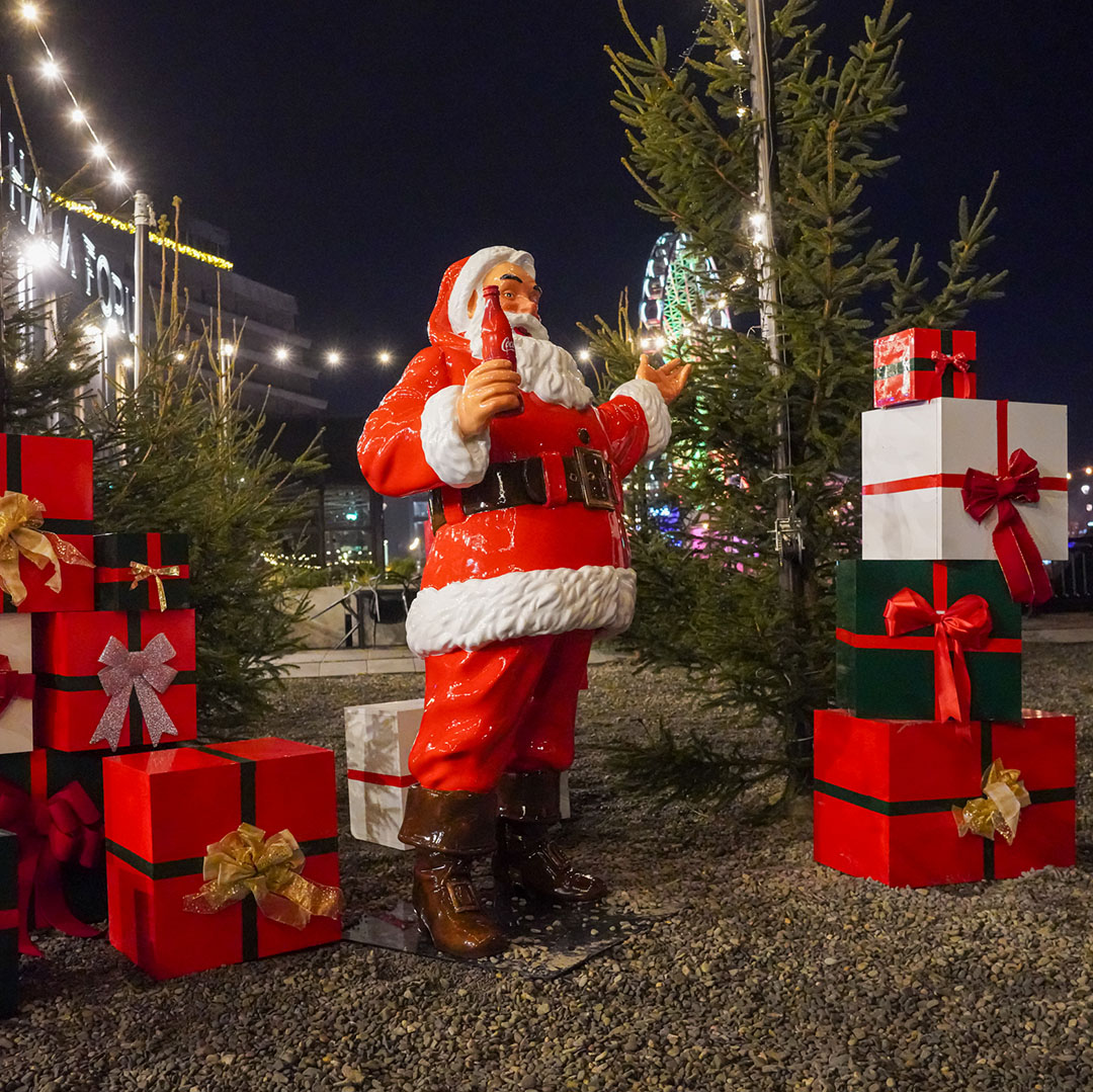 Figurka świętego Mikołaja w czerwonym stroju trzymająca butelkę Coca-Coli, otoczona świątecznymi prezentami z czerwonymi i złotymi kokardami, nocna sceneria z oświetlonymi choinkami i diabelskim młynem w tle