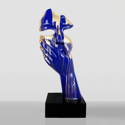 Rzeźba przedstawiająca niepełną, abstrakcyjną twarz trzymaną w dłoni, pomalowana na jaskrawy niebieski kolor z elementami złotego lakieru, umieszczona na czarnej podstawce.